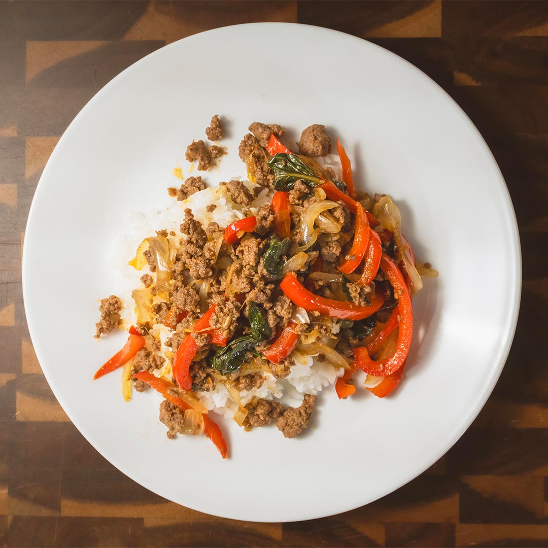 Overhead view of a Thai basil beef stir-fry with sliced red bell peppers and fresh basil over white rice. Thai basil beef bowl with ground beef, red peppers, and basil served over steamed white rice.