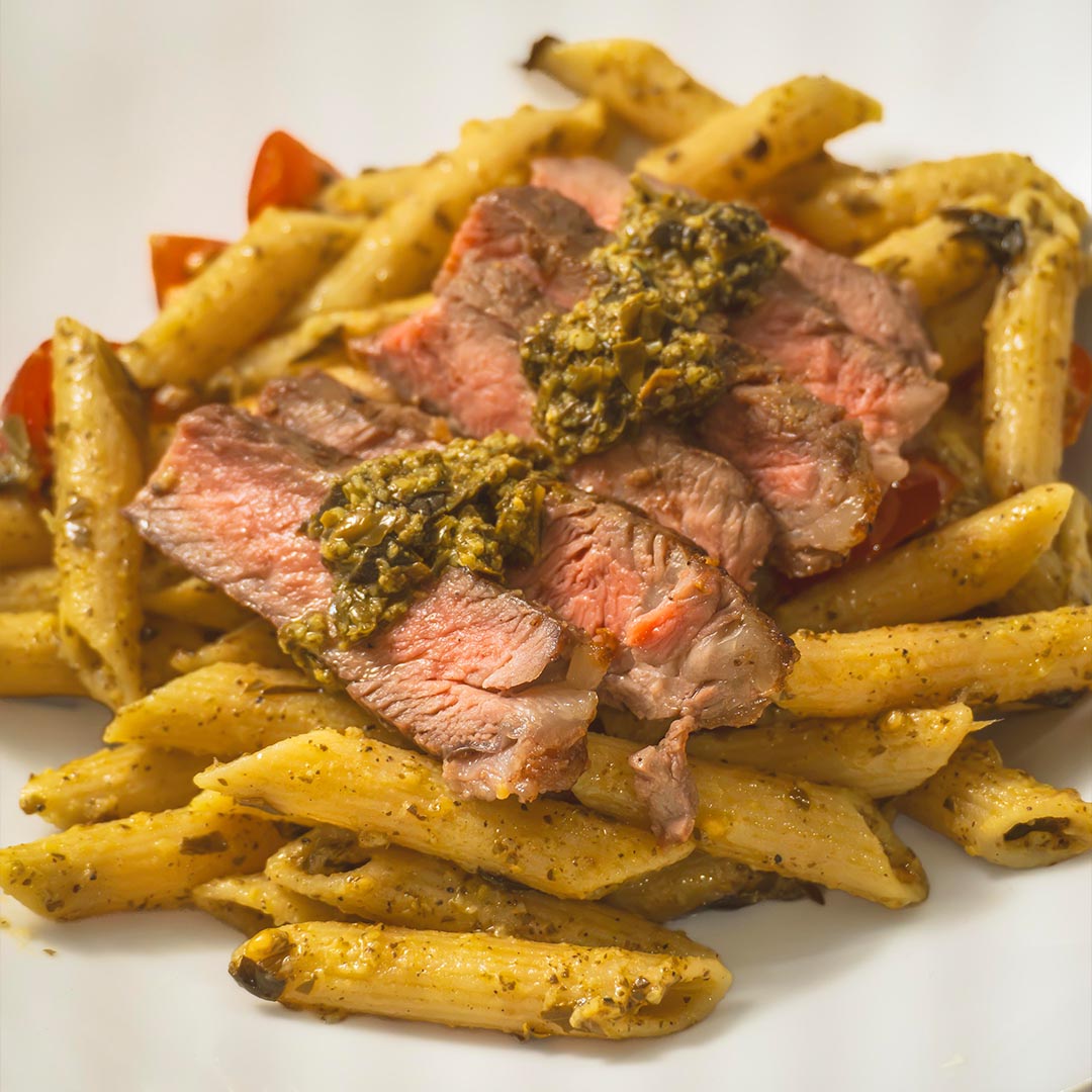 Sliced medium-rare ribeye steak topped with green pesto served over pesto penne pasta and cherry tomatoes