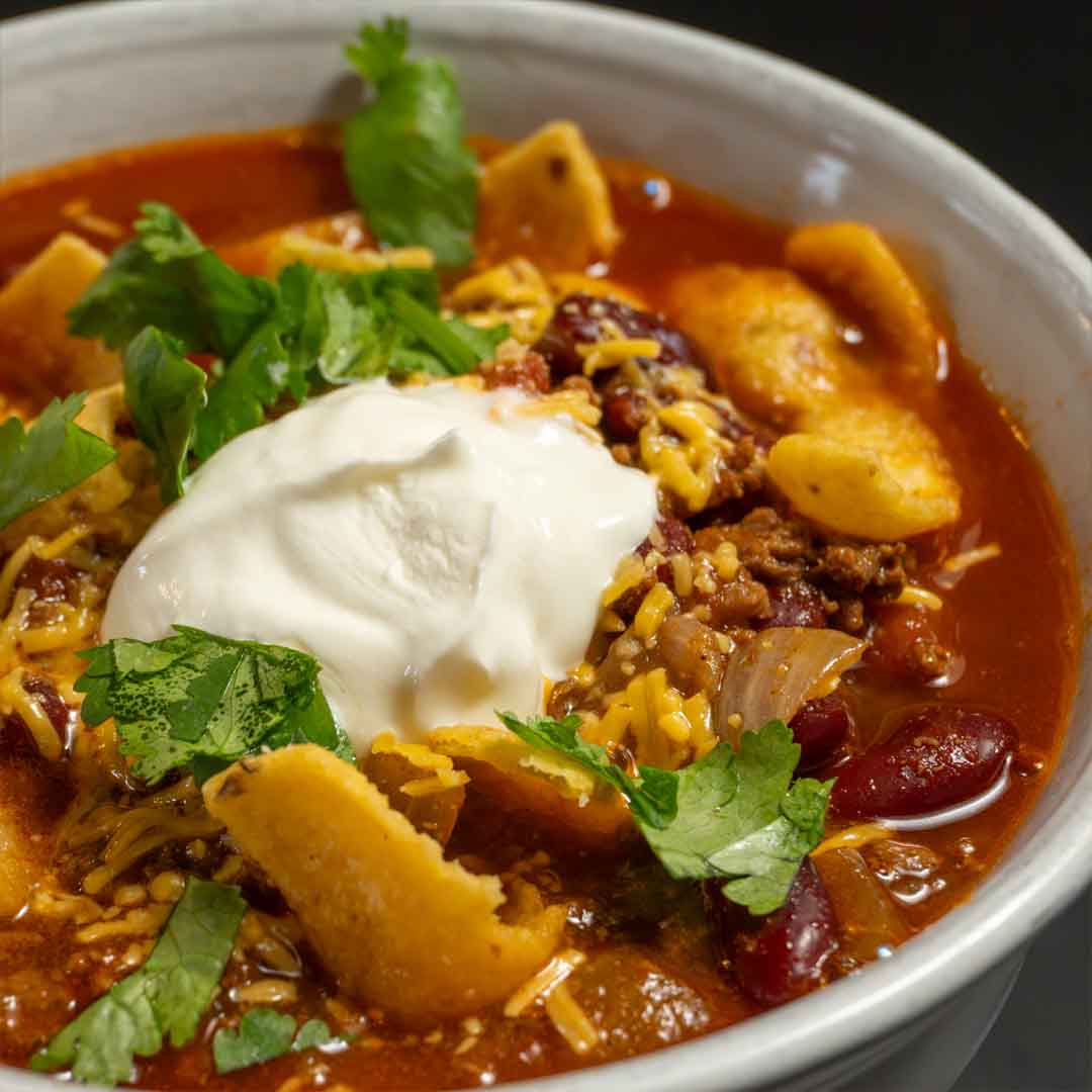 Close-up of a bowl of local beef chili garnished with a scoop of sour cream, melted shredded cheddar, corn chips, and fresh cilantro.