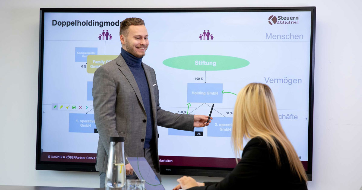 Zertifizierter Stiftungsberater Sven Wecklein präsentiert einer Mandantin ein Doppelholdingmodell mit Stiftung auf einem Bildschirm und erklärt die Struktur der Holding GmbH.