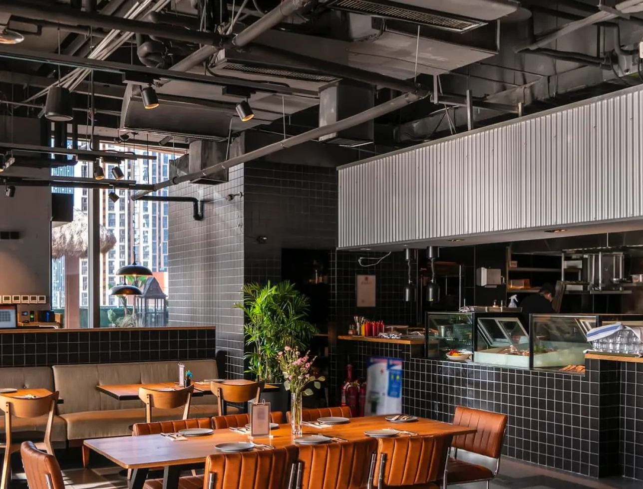 Modern restaurant interior with wooden tables, brown leather chairs, black tiled walls, and a view of the kitchen area.