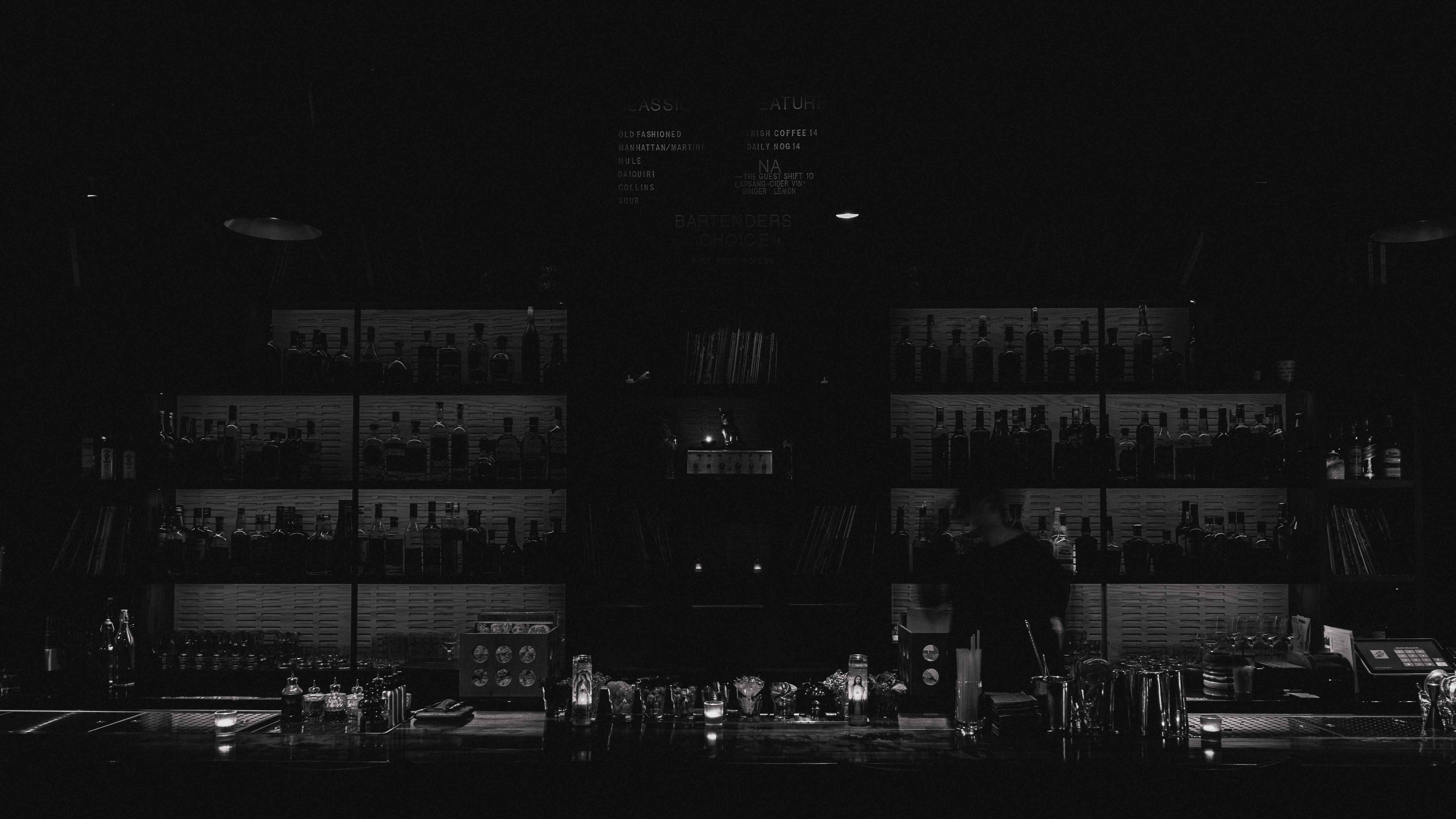 Dimly lit bar with shelves of liquor bottles, candlelit bar counter, and a bartender in motion.
