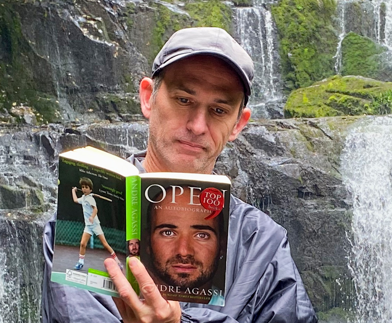 Aaron Elsmore standing in front of a waterfall reading a book.