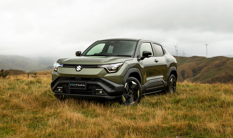 A dark green Suzuki e Vitara all wheel drive vehicle parked on long grass, windmills on hills are visible in the background.