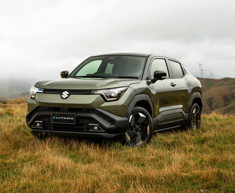 A dark green Suzuki e Vitara all wheel drive vehicle parked on long grass, windmills on hills are visible in the background.