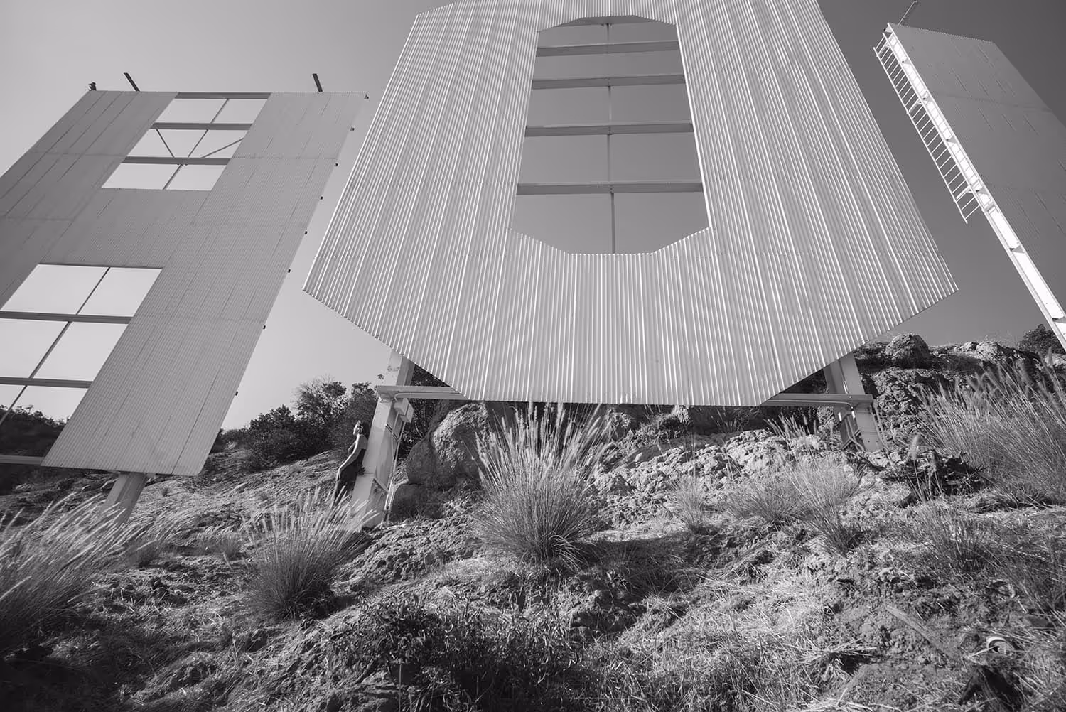 Black and white photo looking up at the back of a large metal sign on a hillside with rough terrain and bushes.