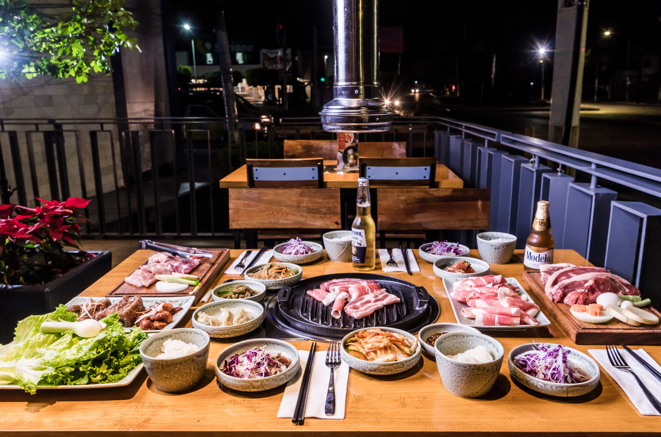 Korean barbecue spread with sliced meats, rice, vegetables, and side dishes surrounding a tabletop grill on an outdoor patio at night.