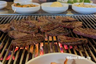 Beef ribs cooking over glowing coals on a tabletop grill, with Korean side dishes in the background at Soot Bull Jip.