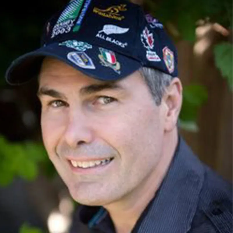 Man wearing a black cap with multiple rugby team patches smiling outdoors.
