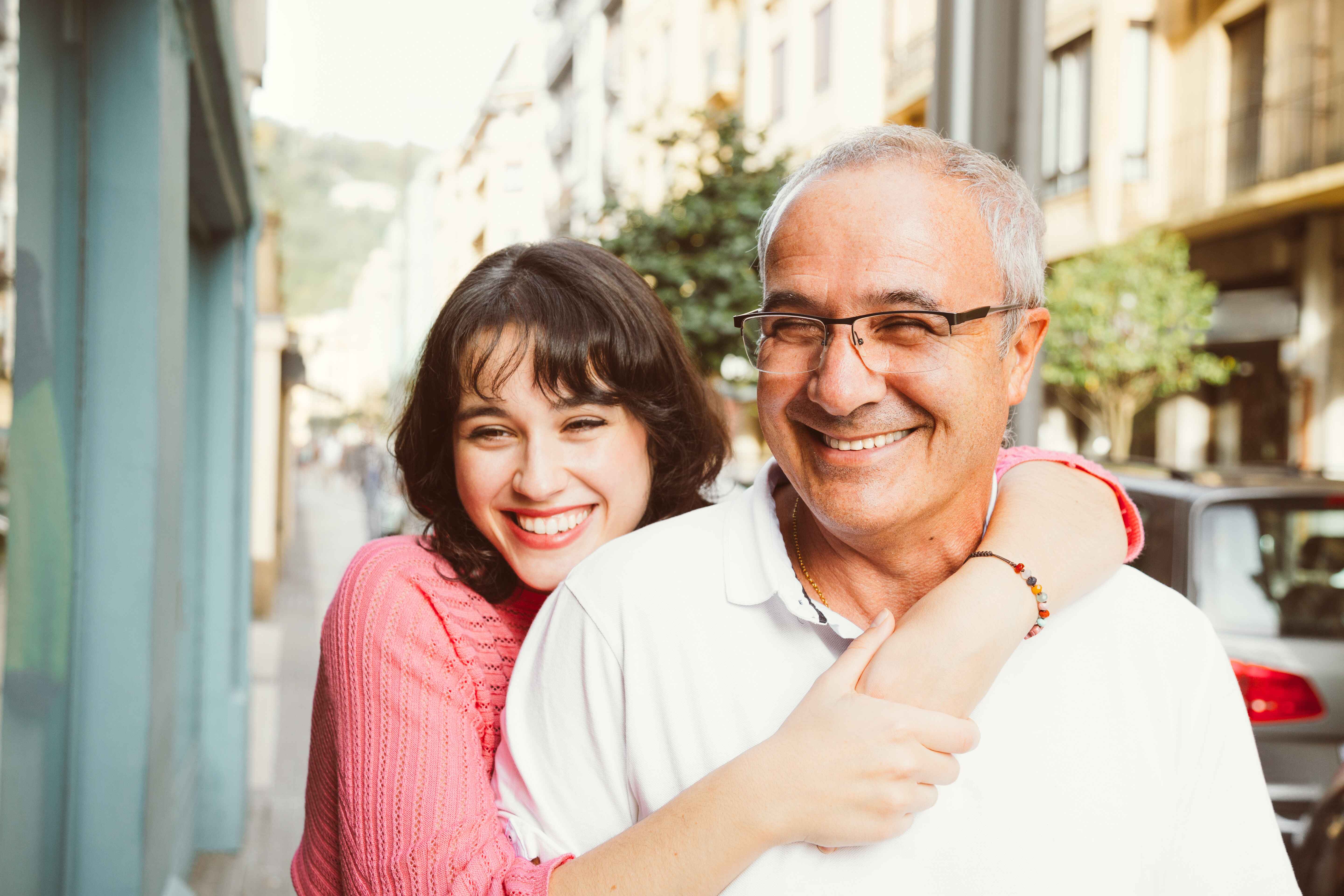 Adult daughter hugging her father from behind on a city street, both smiling