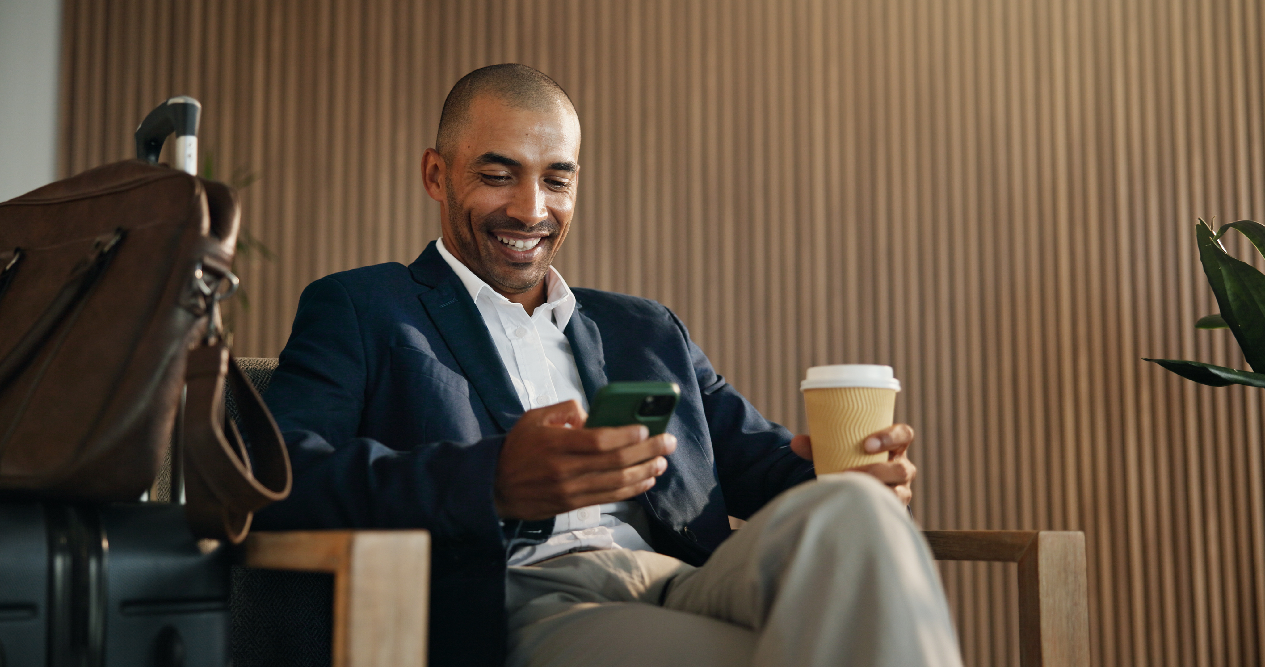 Man in a blazer smiling at his phone while holding a coffee cup in a hotel lounge