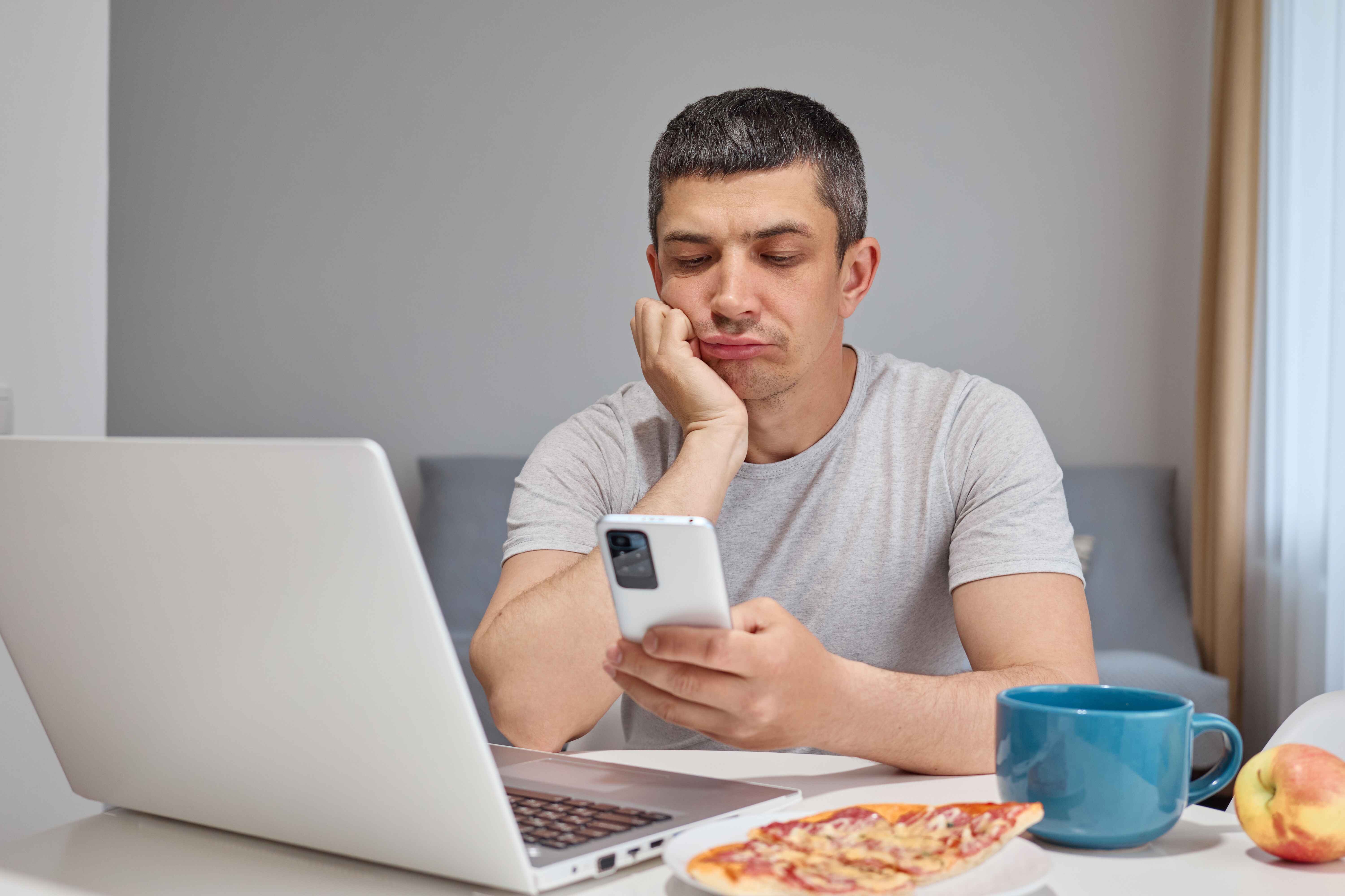 Man looking frustrated while using a dating app on his phone beside a laptop