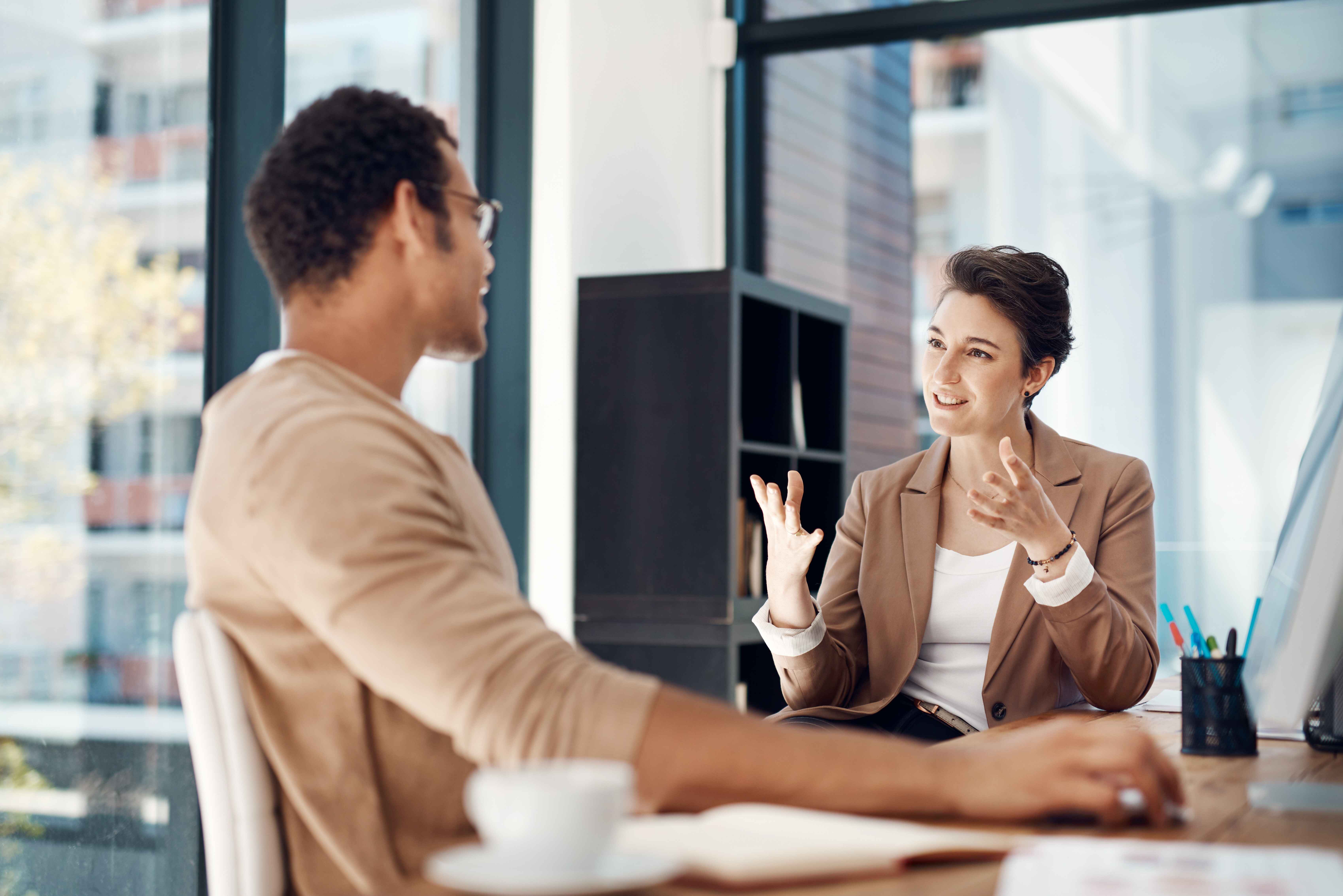 Man and woman having an animated conversation at an office desk, woman gesturing with her hands