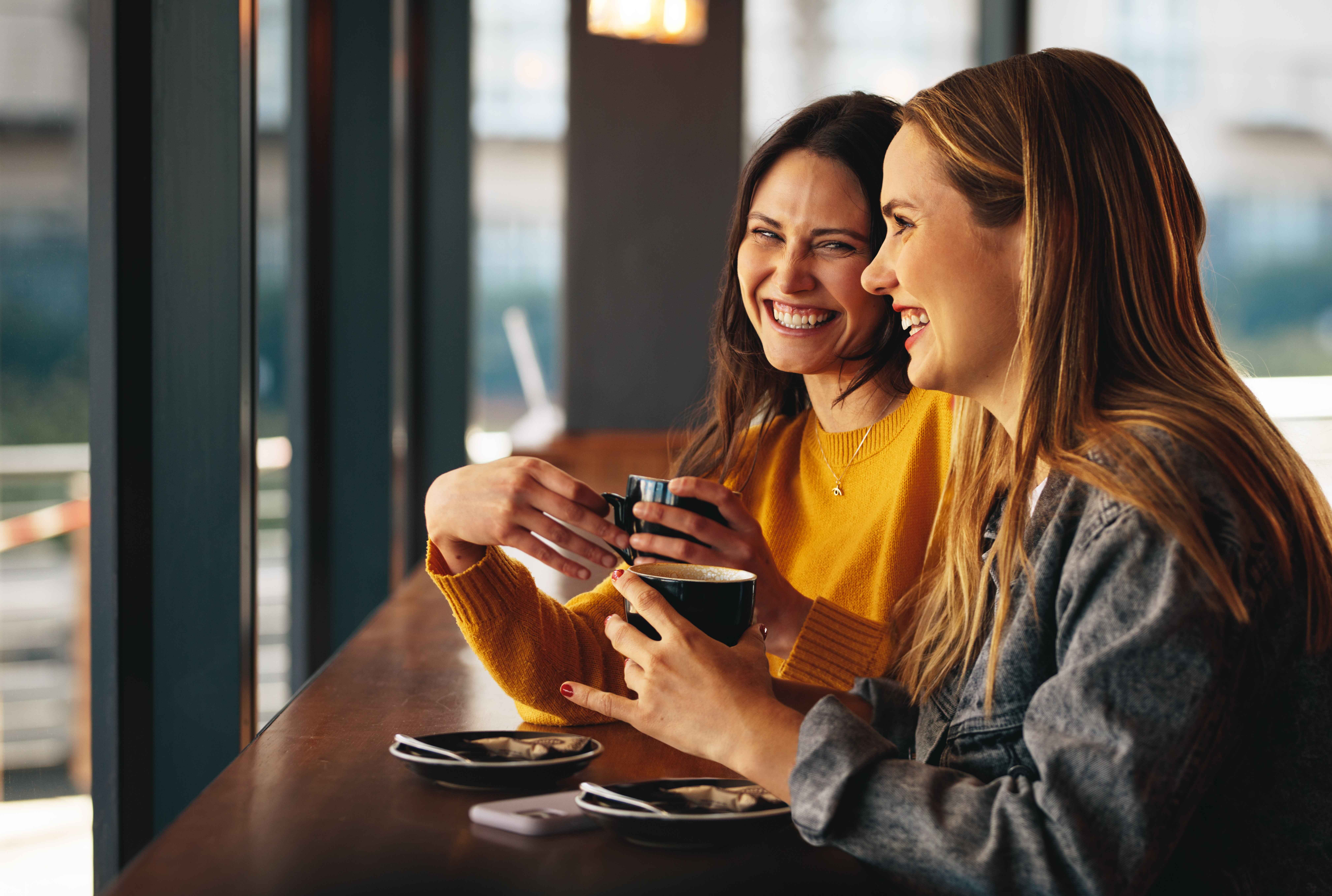 Two friends laugh together over coffee at a sunny café table.