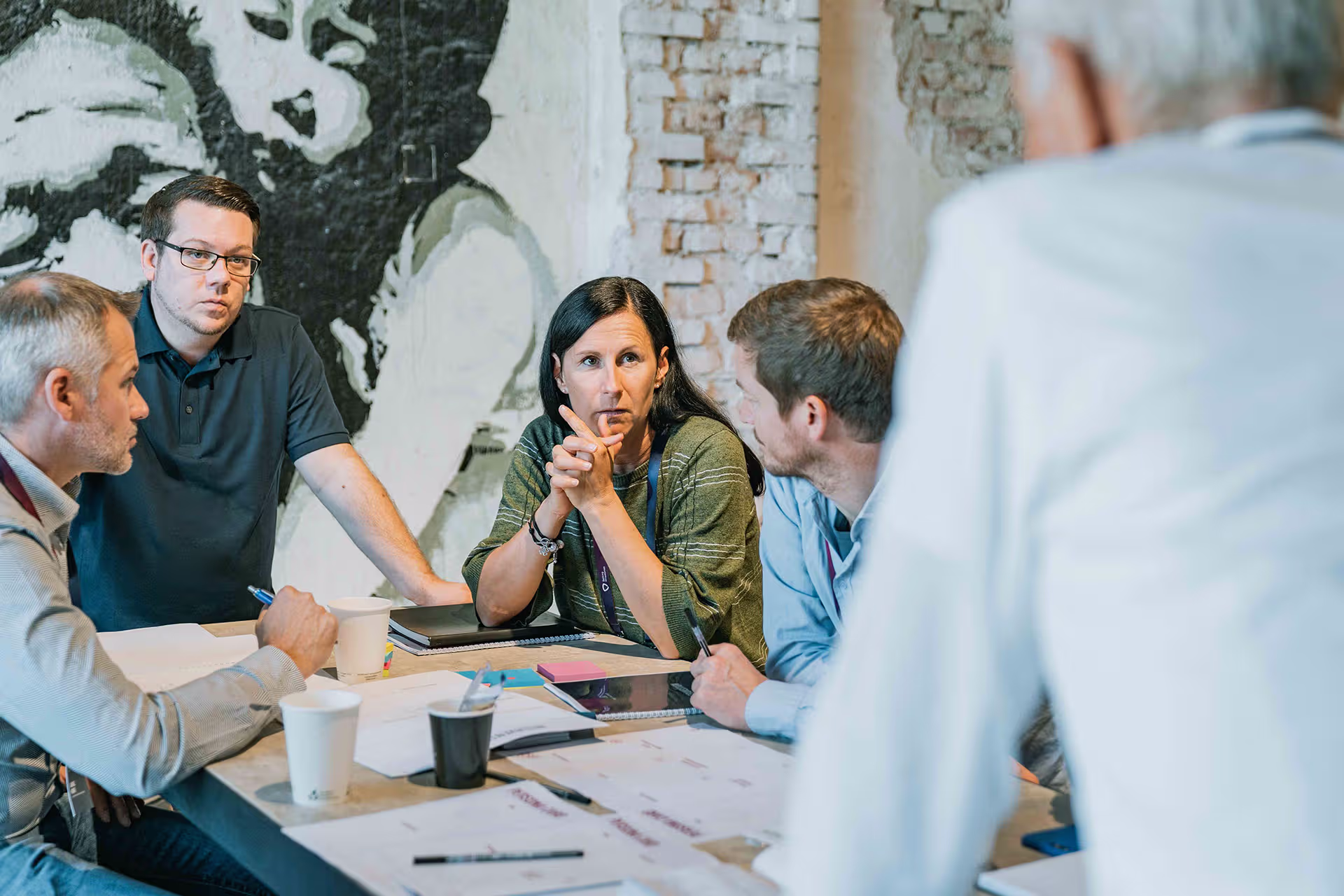Fünf Personen diskutieren konzentriert an einem Tisch mit Dokumenten vor raue Backstein- und Wandmalerei-Hintergrund.