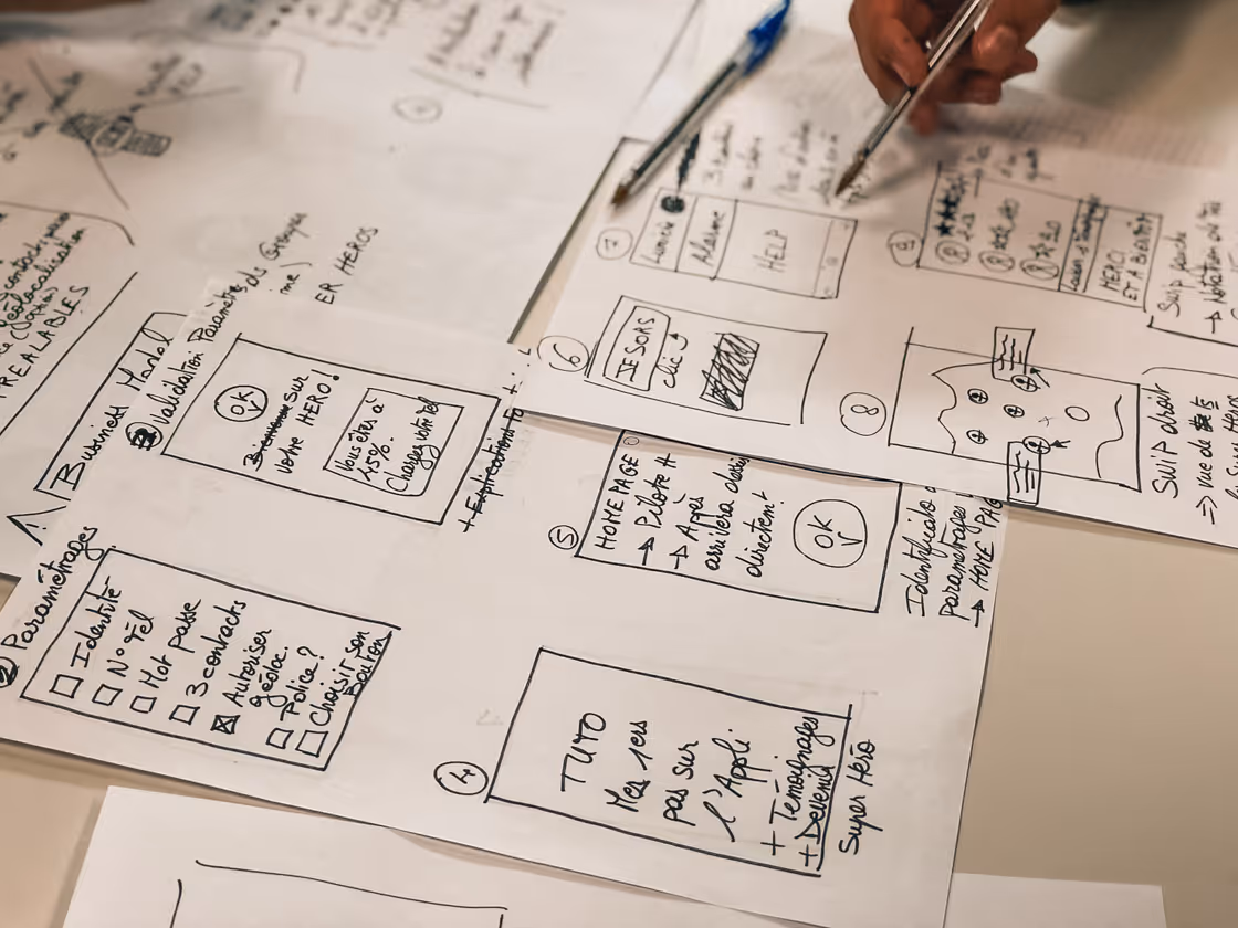 Nahaufnahme von mehreren handgezeichneten Papier-Layouts mit Notizen und Skizzen für eine App- oder Webseitenstruktur, während jemand mit einem Stift darauf zeigt.
