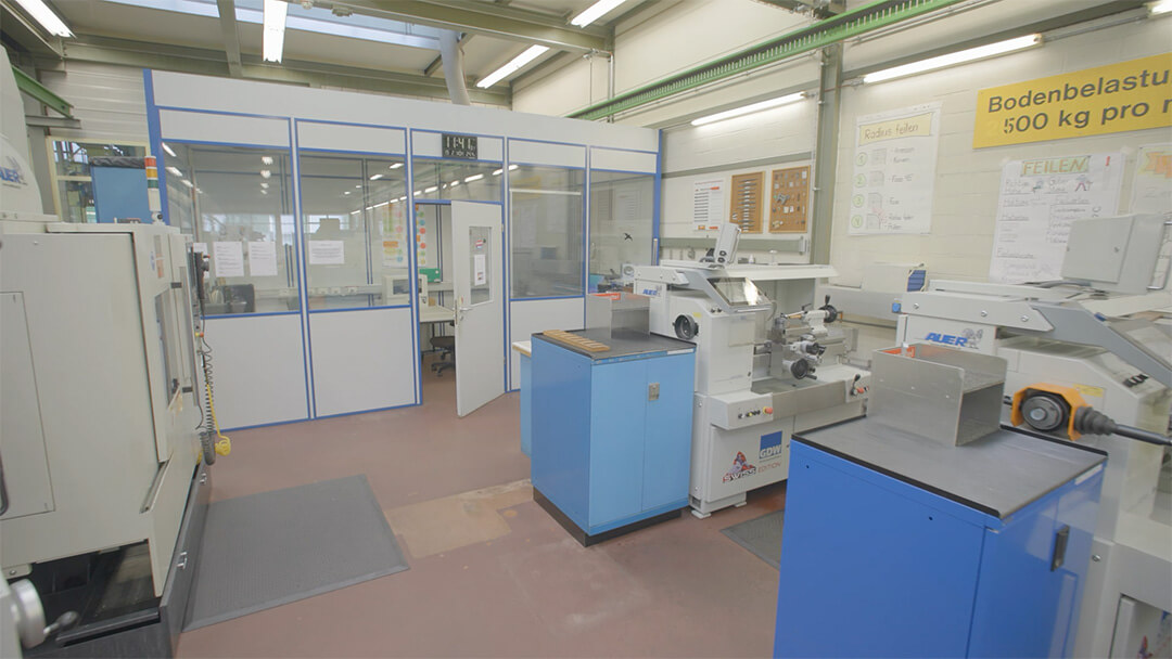 Werkstatt mit Metallbearbeitungsmaschinen und blauen Schränken, Bürozimmer mit Glastüren hinten.