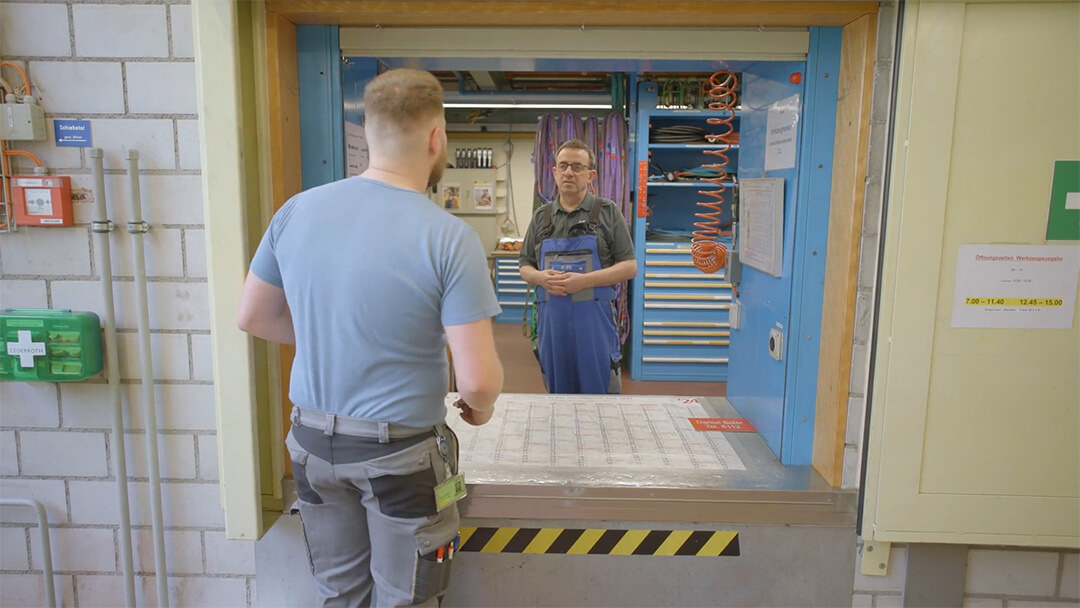 Werkzeugausgabe. Zwei Männer sprechen an einem Industriearbeitsplatz mit Werkzeugeinrichtung im Hintergrund.