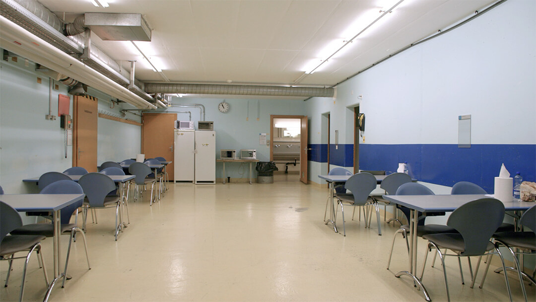 Kantine mit blau-grauen Stühlen, Tischen, Kühlschränken und Mikrowellen in einem Raum mit hellblauen Wänden.