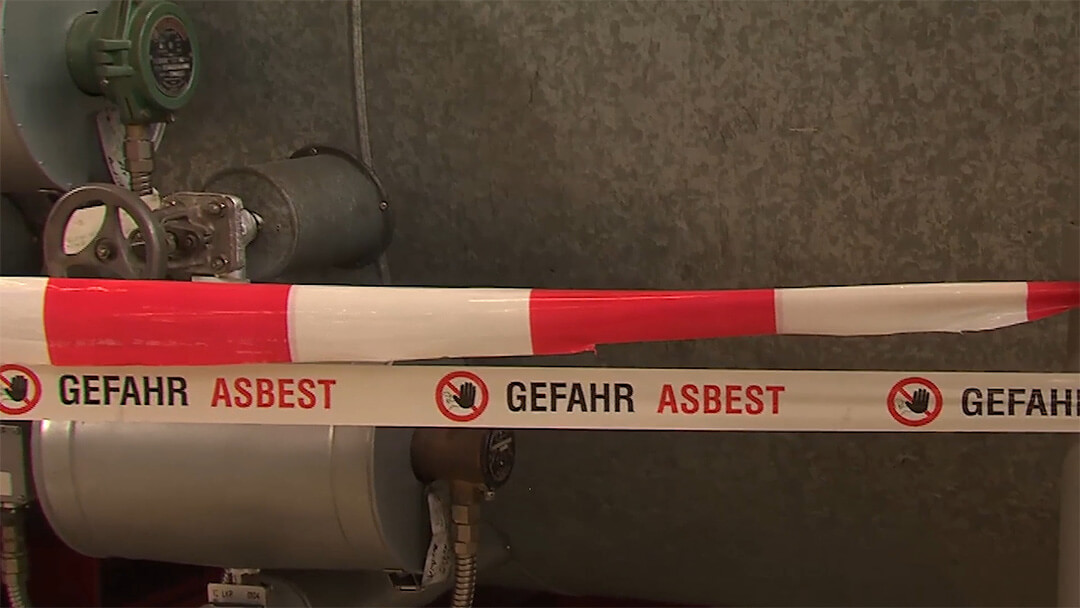 Absperrband mit der Aufschrift Gefahr Asbest vor Metallrohren und Ventilen.