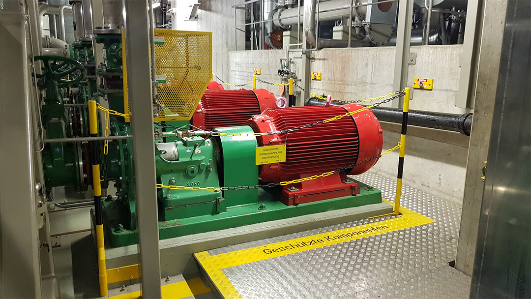 Industriemotor mit rotem Gehäuse und grünem Sockel, abgesperrt mit Ketten und Warnhinweis.