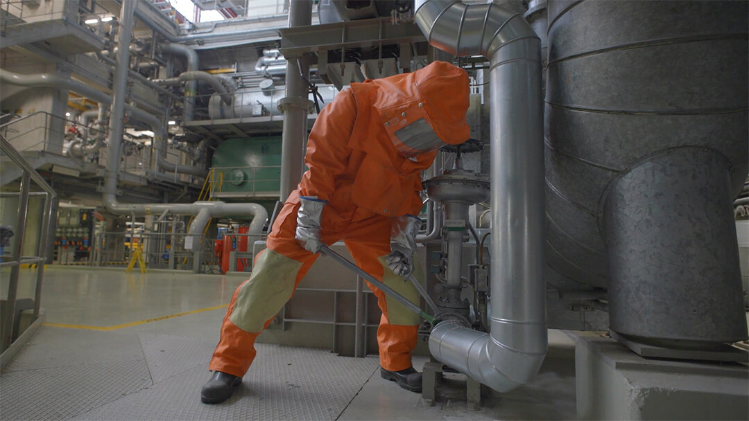 Eine Person in einem orangefarbenen Schutzanzug, die in einer Industrieanlage mit Rohrleitungen ein Ventil einstellt.