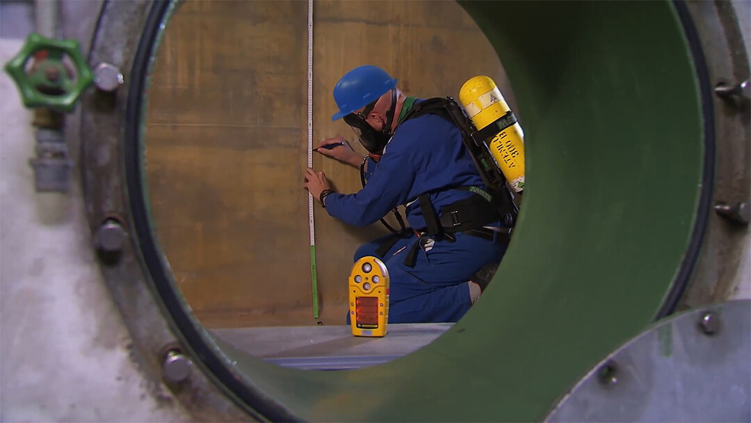 Techniker mit Schutzhelm und Atemschutzgerät misst Wandhöhe in einem technischen Wartungsraum.