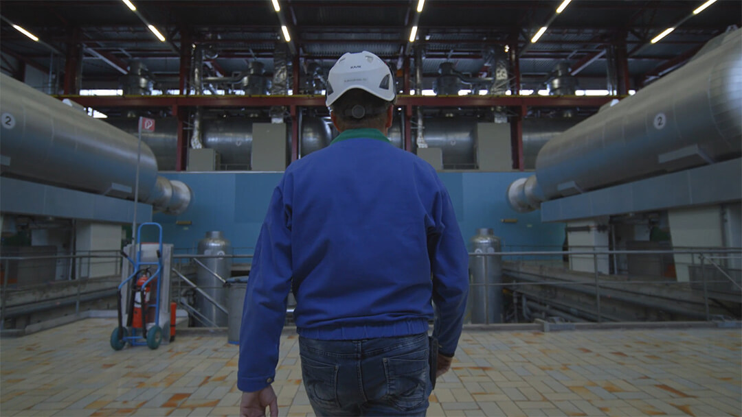 Arbeiter mit weissem Helm und blauer Arbeitsjacke in einer grossen Industrieanlage.