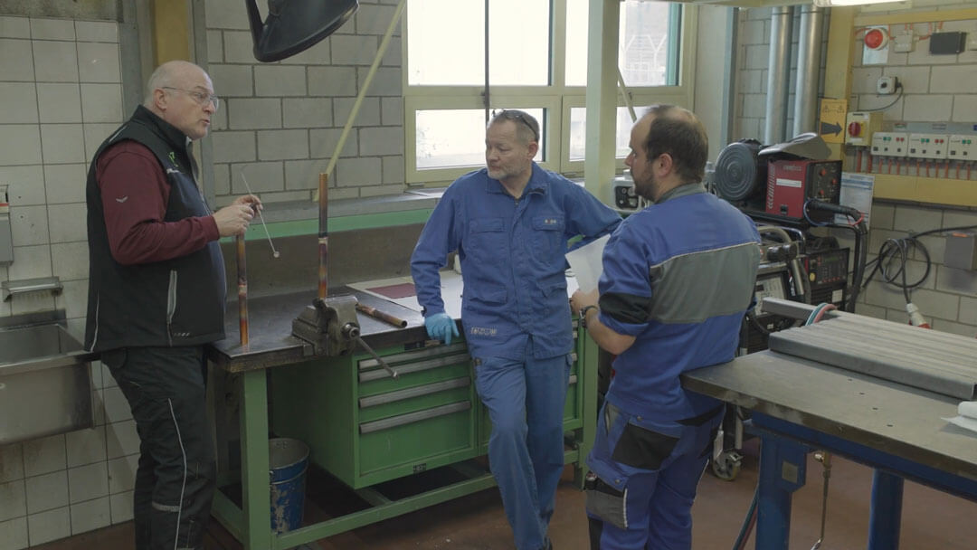 Drei Männer reden in einer Werkstatt mit Werkzeugen auf einem grünen Tisch und Maschinen im Hintergrund.