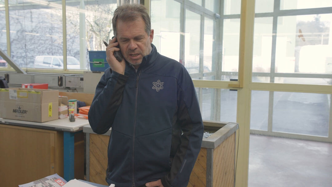 Mann in blauer Jacke telefoniert in einem Büro mit Kartons und Fenster im Hintergrund.