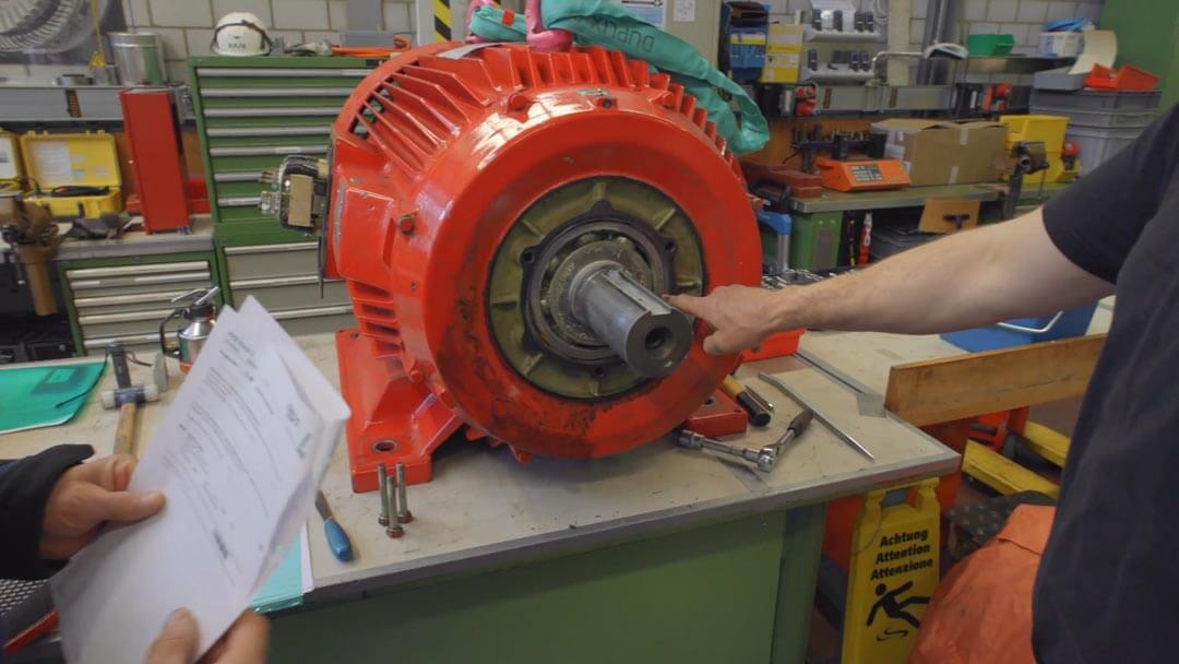Person zeigt auf die Welle eines grossen roten Elektromotors auf einer Werkbank.