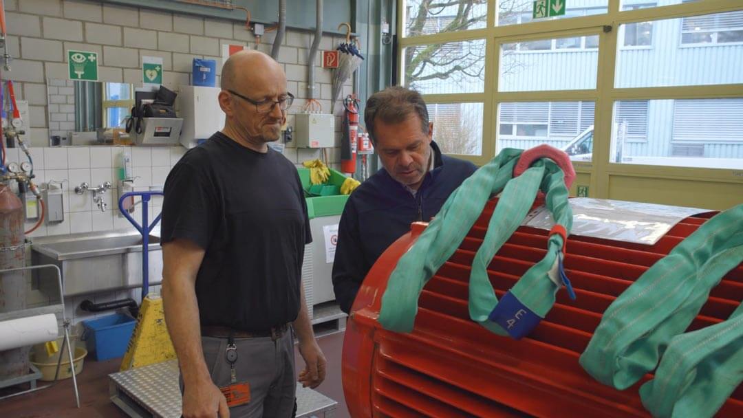Zwei Männer begutachten eine große rote Industrieausrüstung mit grünen Hebebändern in Werkstatt.