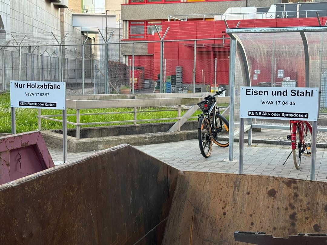 Abfallcontainer für Holzabfälle und einen für Eisen und Stahl vor einem Gebäude mit Fahrrädern.