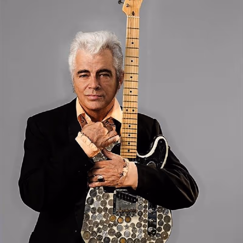 A well-dressed man with gray hair holds an electric guitar in front of a plain gray background.