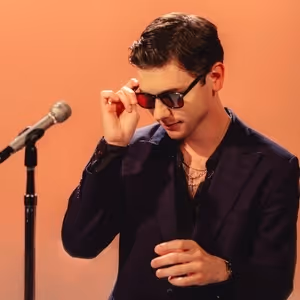 A man wearing a dark suit and sunglasses stands at a microphone, performing on a stage with an orange background.