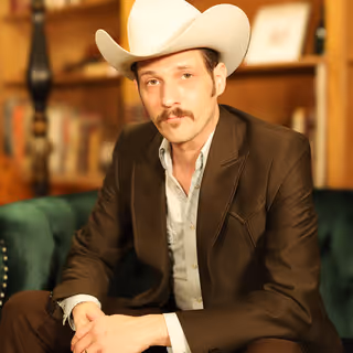 A man wearing a cowboy hat and a brown suit is sitting in a room with a green chair and other furniture in the background.