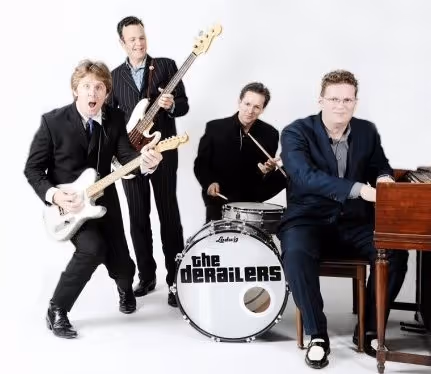 A group of four men, dressed in suits, performing as a rock band with electric guitars, drums, and a piano.
