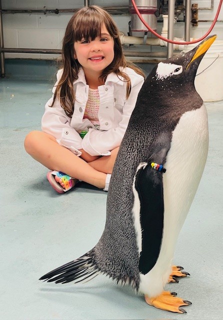 A young girl with long brown hair sits on the floor next to a large penguin, both appearing to be in an indoor setting with a tiled floor and equipment in the background.