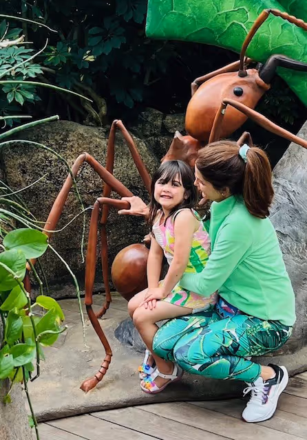 Two young children, a girl and a boy, are sitting on the ground in a lush, green outdoor environment, examining a large, wooden insect sculpture.