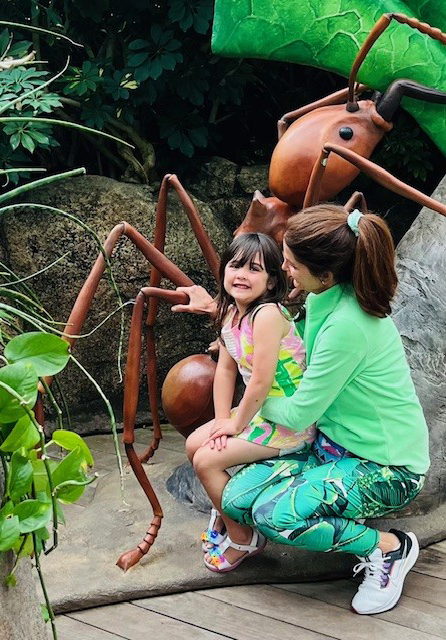 Two young children, a girl and a boy, are sitting on the ground in a lush, green outdoor environment, examining a large, wooden insect sculpture.