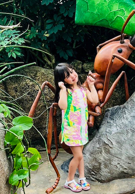A young girl in a colorful dress stands in a lush, green garden surrounded by large leaves and a giant wooden insect sculpture.
