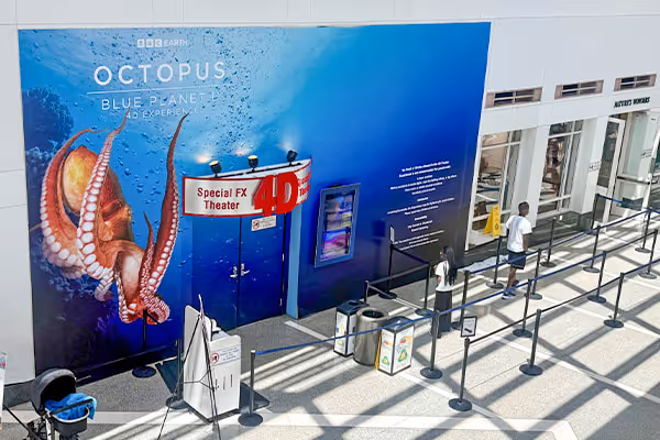 The image depicts a large blue and red banner advertising the "Octopus: Blue Planet" exhibit, with a giant octopus illustration in the center. In the foreground, there are several stanchions and barriers, suggesting this is a public exhibition or event space.

