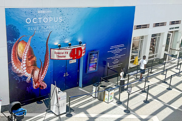 The image depicts a large blue and red banner advertising the "Octopus: Blue Planet" exhibit, with a giant octopus illustration in the center. In the foreground, there are several stanchions and barriers, suggesting this is a public exhibition or event space.

