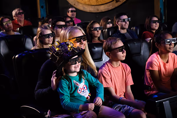 A group of people, mostly children, wearing 3D glasses and sitting in a dark theater, with a large screen in the background.

