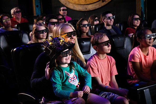A group of people, mostly children, wearing 3D glasses and sitting in a dark theater, with a large screen in the background.

