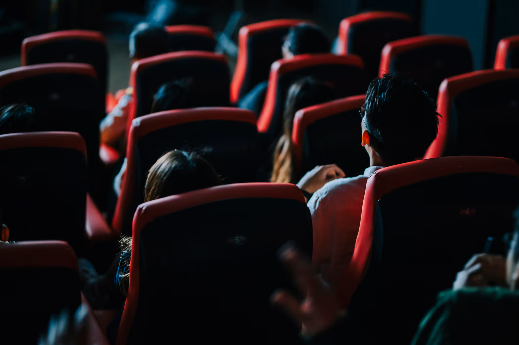 Rows of red theater seats fill the frame, with the dim lighting creating a sense of anticipation in the darkened auditorium.

