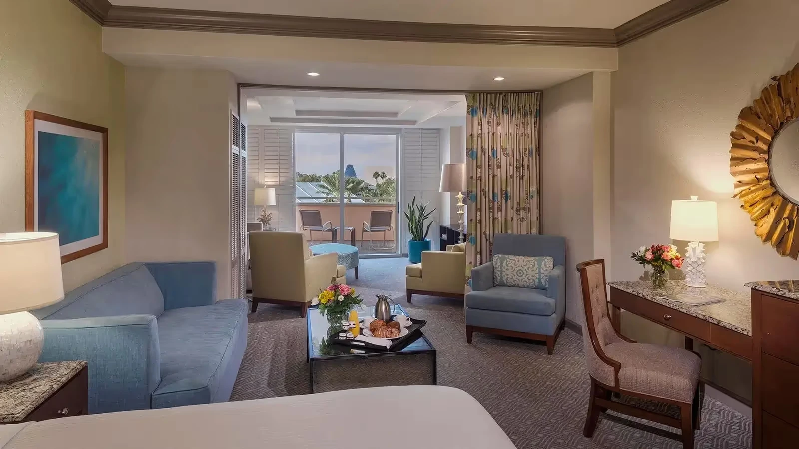 Hotel room with seating area including blue and beige armchairs, a blue sofa, a desk with a chair, and glass doors leading to a balcony with outdoor chairs.
