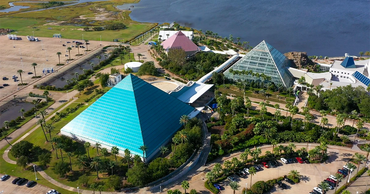 An aerial view of a colorful and vibrant theme park with various architectural structures, including pyramids and other unique buildings, surrounded by lush greenery and a body of water in the background.