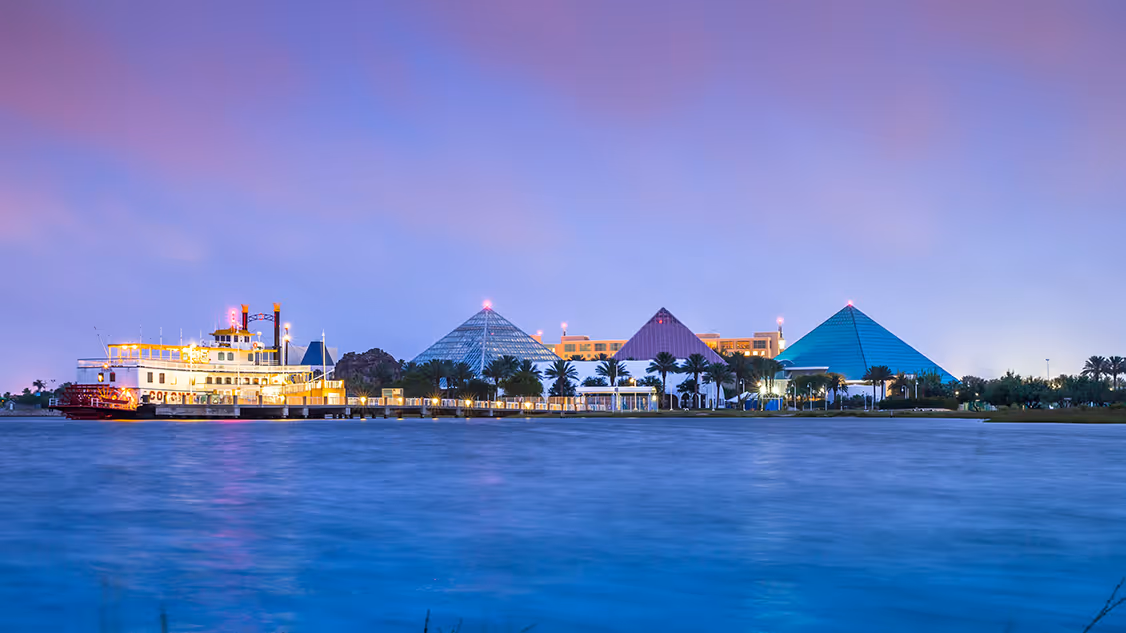 "Moody Gardens Hotel, Attractions & Golf Course in Galveston Island, Texas, featuring the iconic glass pyramids and the Colonel Paddlewheel Boat illuminated at dusk, viewed across the water with a vibrant evening sky.