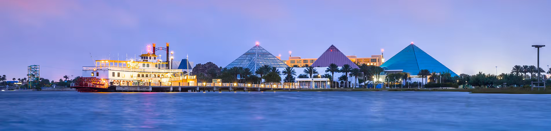 A vibrant cityscape with colorful, pyramid-shaped buildings along the waterfront, surrounded by palm trees and a serene body of water reflecting the illuminated skyline at Moody Gardens in Galveston, Texas 